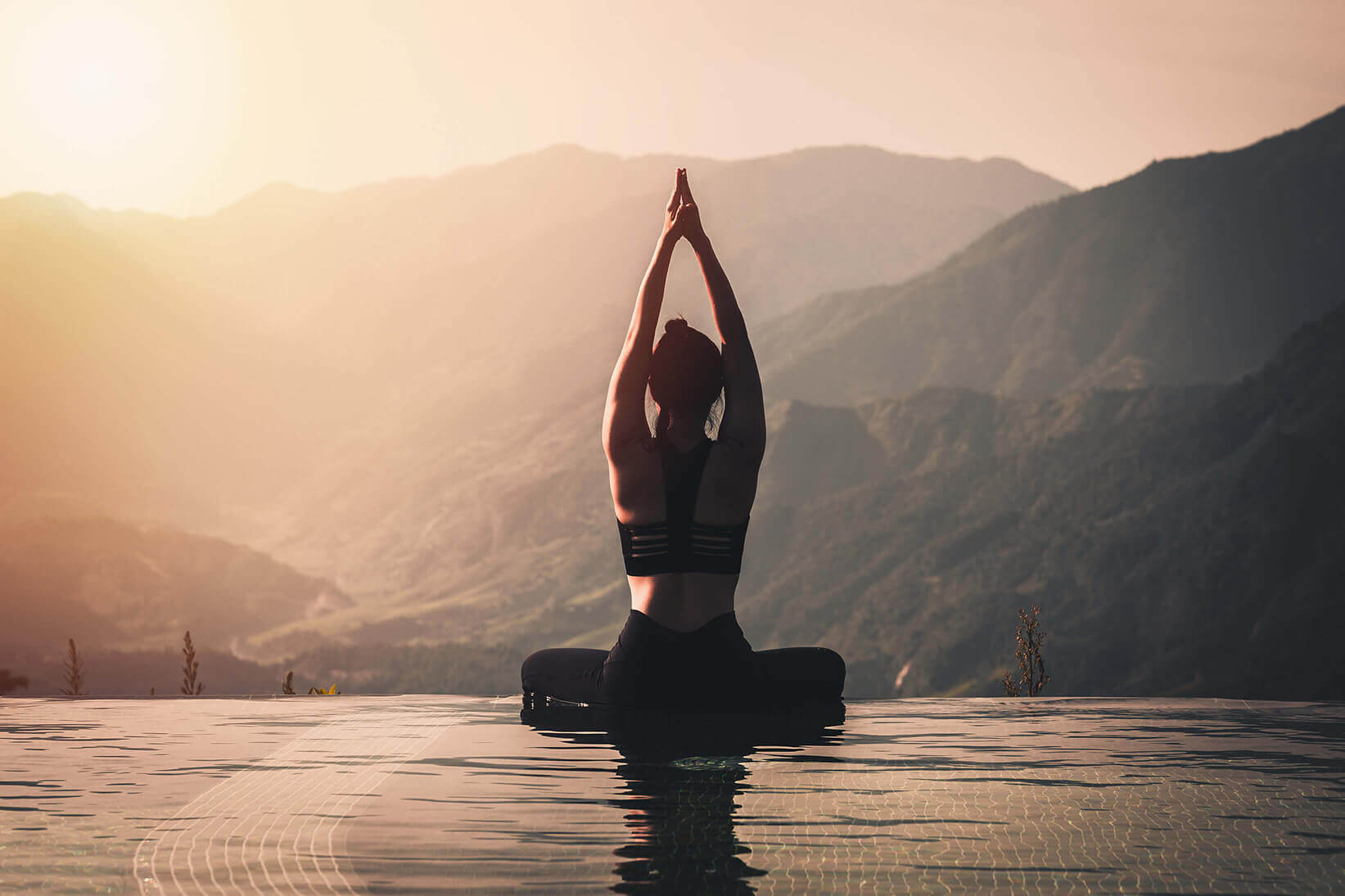 A person sitting cross legged with hand over their head with a mountain view and sunset