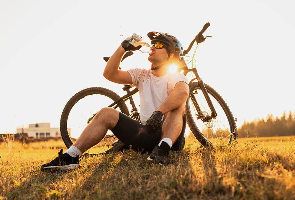 A man outdoors, leaning against his bicycle while drinking from his water bottle. 